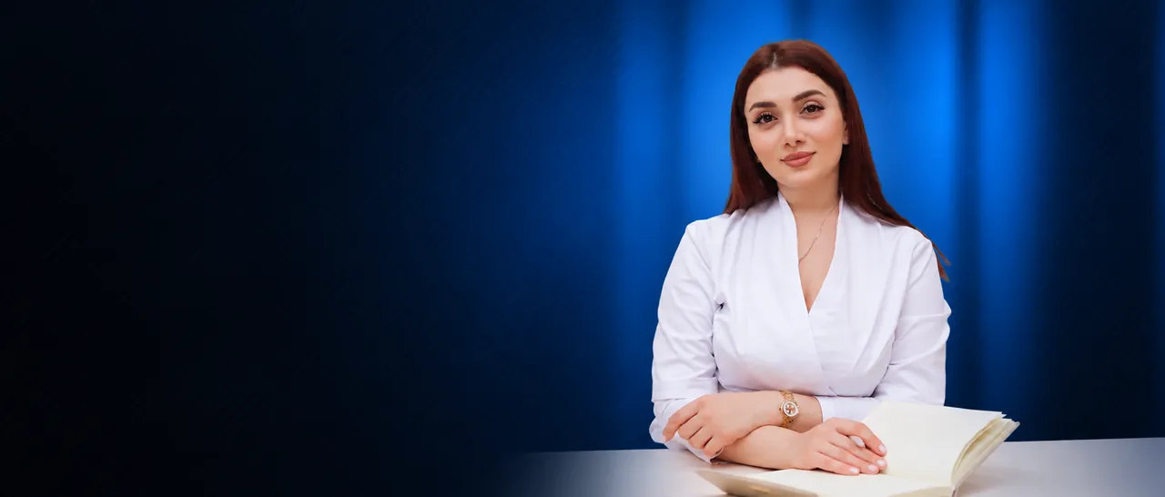 Dr. Anjela in a white coat seated at a desk with an open notebook, blue backdrop