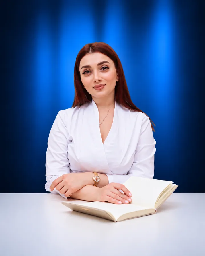 Dr. Anjela in a white coat seated at a desk with an open notebook, blue backdrop